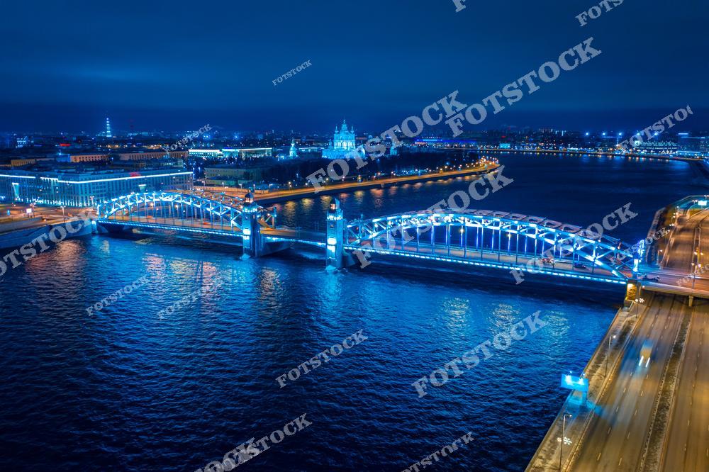 На переднем плане изображен Большеохтинский мост, который соединяет исторический центр Санкт-Петербурга с районом Охта. В ночное время мост красиво подсвечен, его металлические конструкции отражаются в воде Невы. На заднем плане виден Смольный собор, построенный по проекту Бартоломео Растрелли в XVIII веке, который является одним из архитектурных символов города.